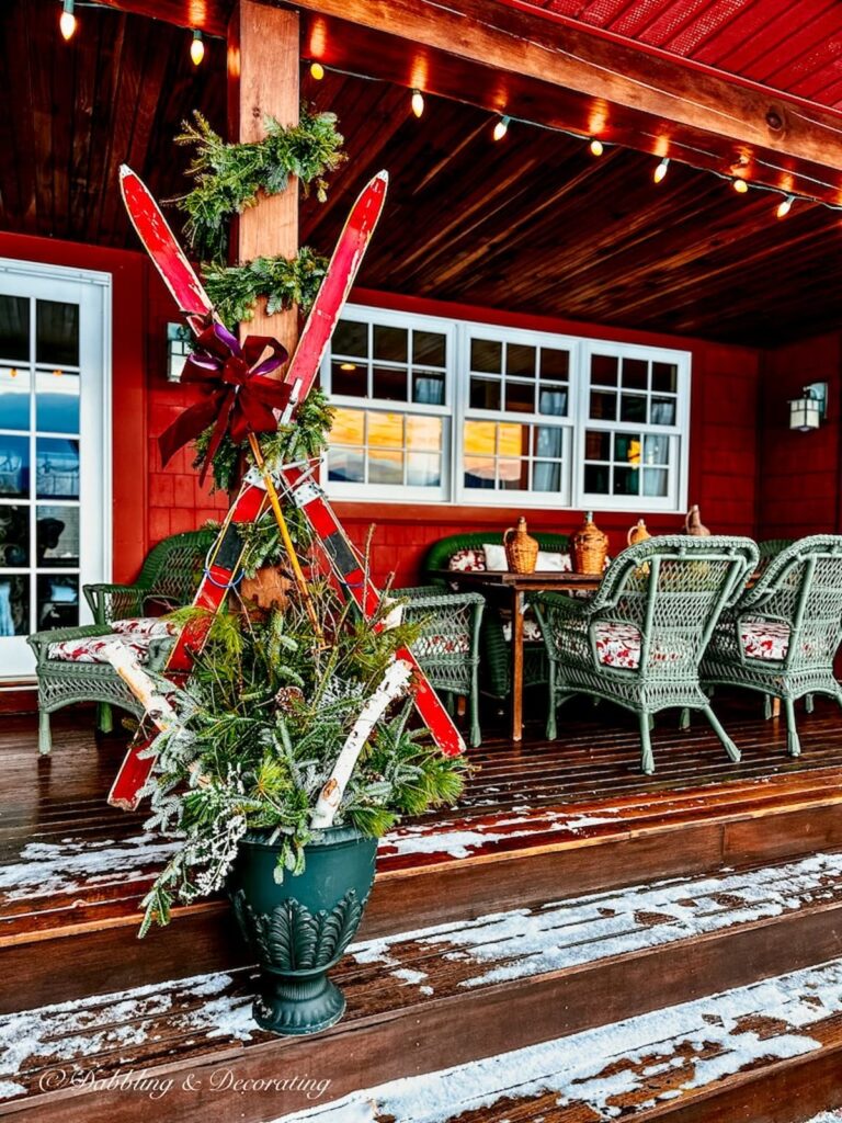 Vintage Christmas red skis hanging on porch with evergreen garland and urn filled with evergreens and birch logs on porch with Christmas lights.