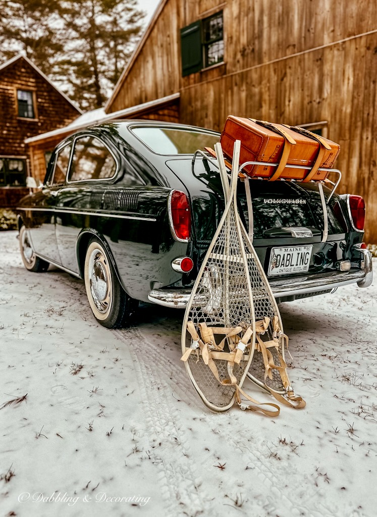 Vintage green VW Fastback with white vintage snowshoes in the snow with vintage leather suitcase attached to trunk in front of rustic cedar shakes home.