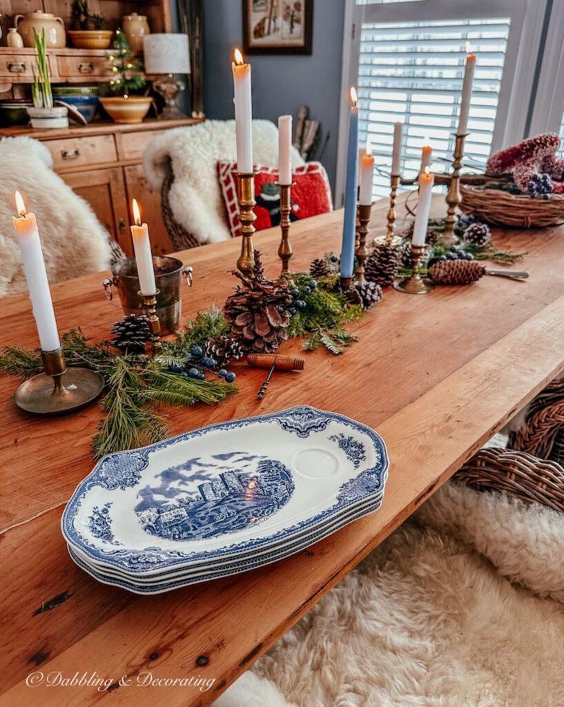 Several vintage brass candlesticks lined up on pine table with greenery, pinecones, and vintage blue tea and toast plates.