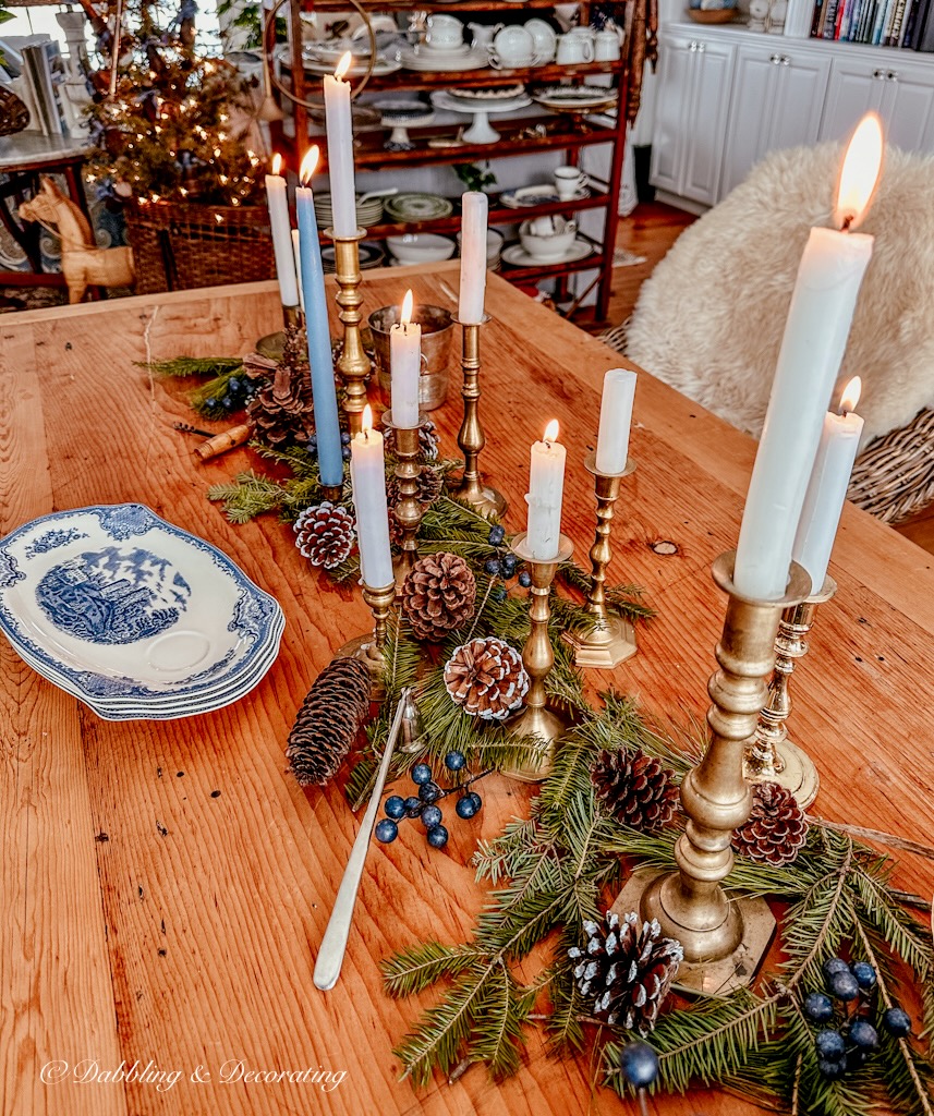 Several vintage brass candlesticks lined up on pine table with greenery, pinecones, and vintage blue tea and toast plates.