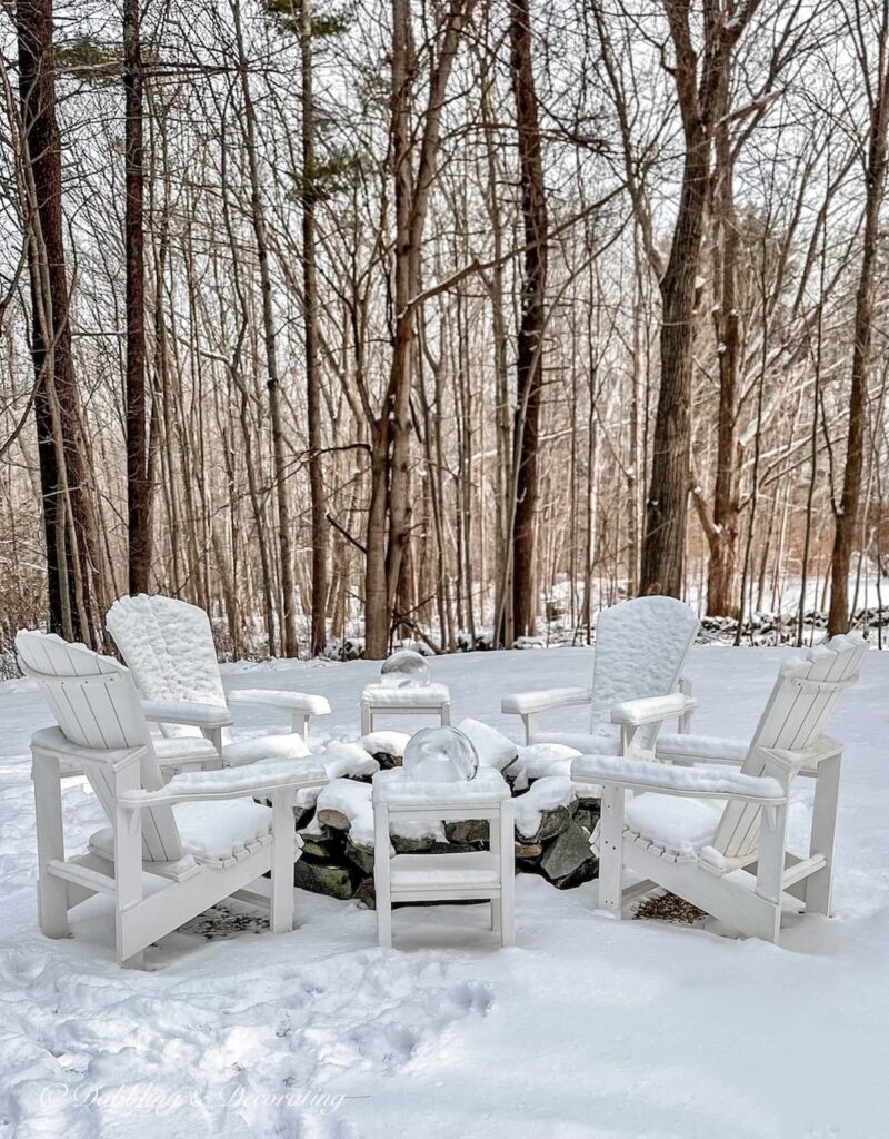 Outdoor stone fire pit surrounded by white Adirondack chairs in the snow.