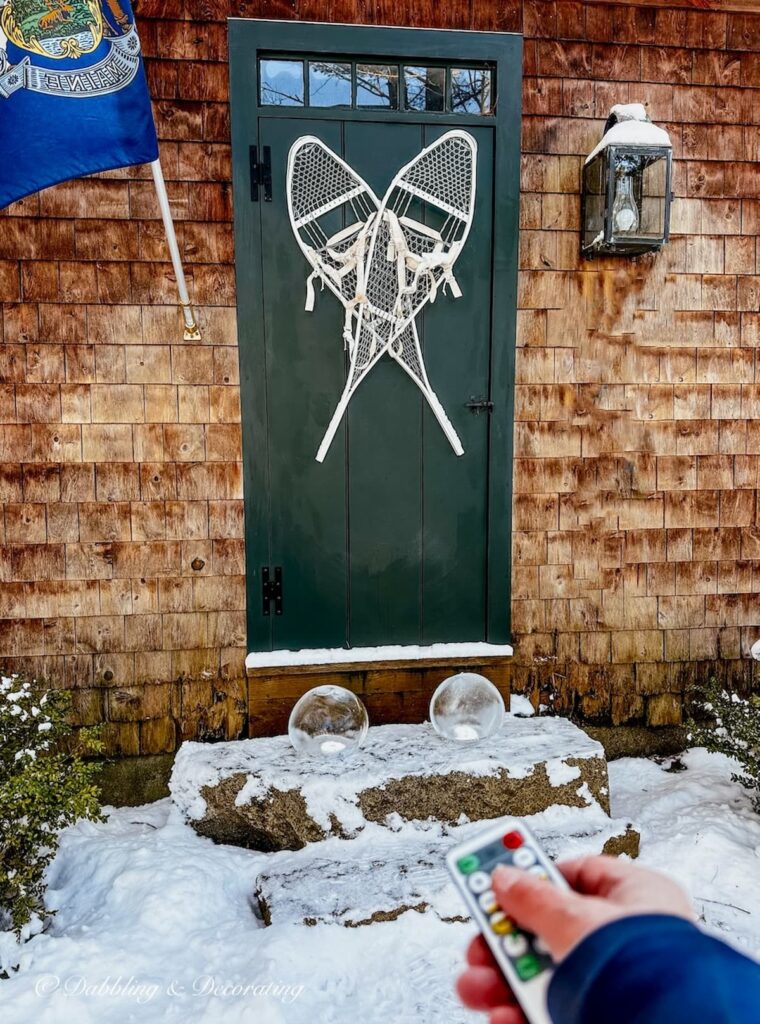 Cedar shake home with dark green front door with white snowshoes with two ice lanterns on door step and Maine flag.