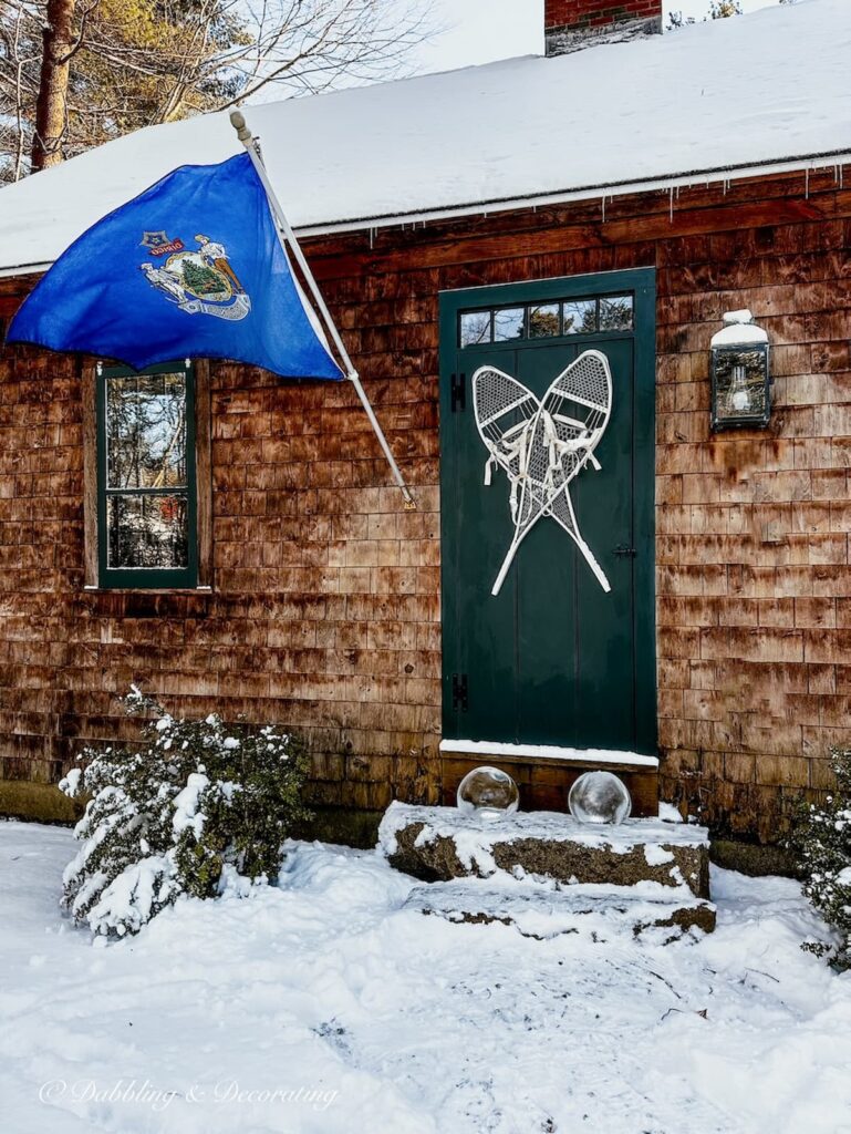 White snowshoes on green front door of rustic cedar shakes home with ice lanterns on front step and Maine flag in snow.