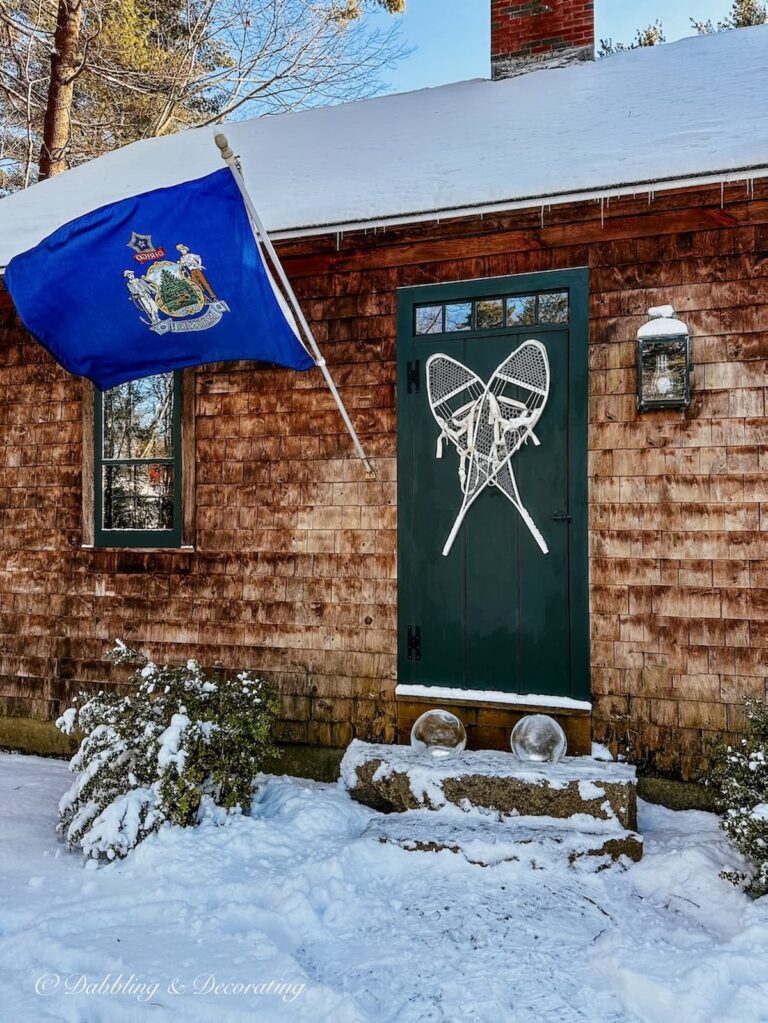 Cedar Shakes house with Maine flag at front door with snowshoes and ice lanterns on snowy steps.