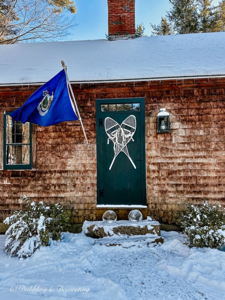 Ice lantern globes on stone steps in front of door with snowshoes on cedar shakes home with Maine flag.