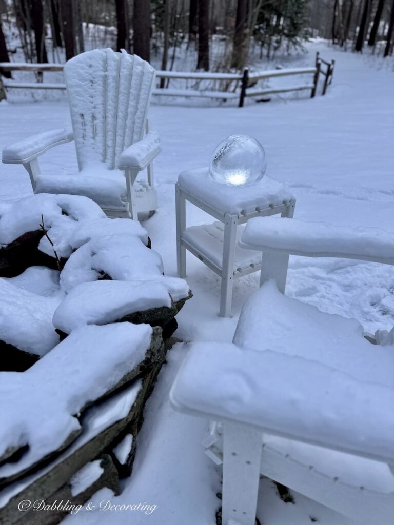 White Adirondack chairs with lit ice lantern in the snow around stone fire pit.