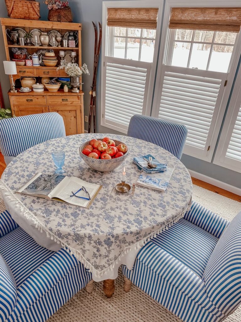 Blue and white dining room table with vintage hutch and three large windows with bamboo shades and blinds.