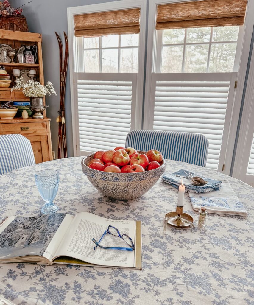 Bowl of apples on blue and white dining room table with three windows with bamboo shades and vintage hutch.