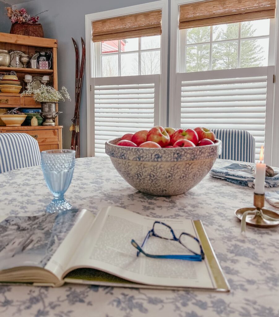 Bowl of apples on blue styled dining room table with bamboo shades on windows with white blinds.