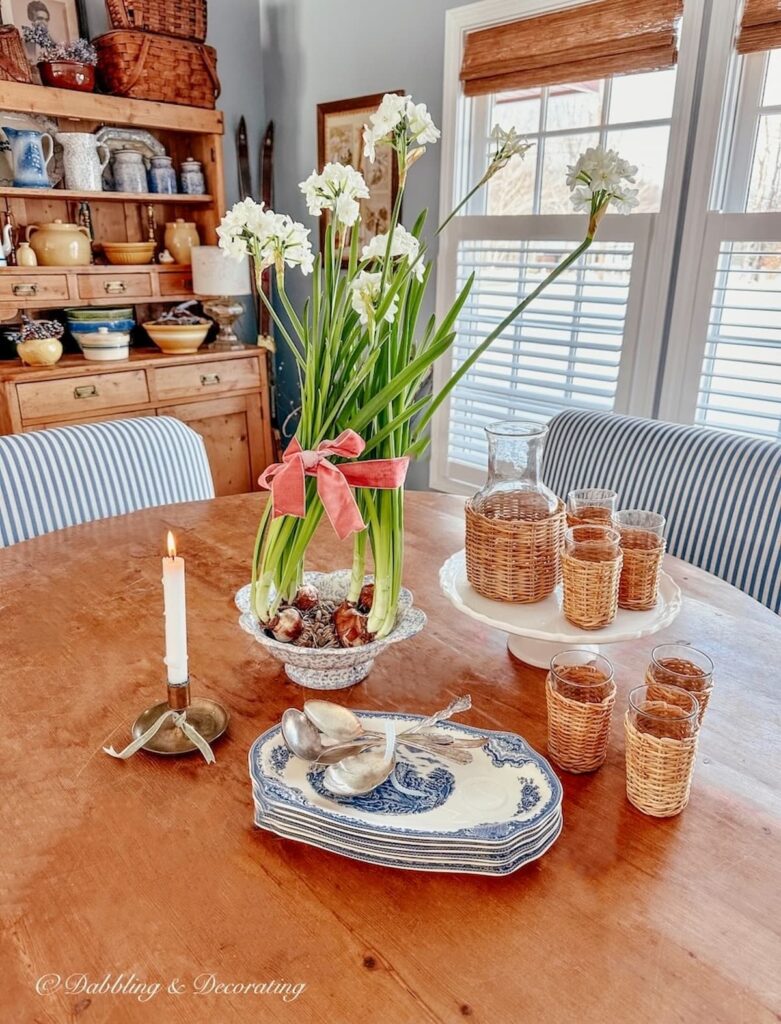 Paperwhites and vintage dishes with candle on pine dining room table, hutch and bamboo shades and shutters on windows.