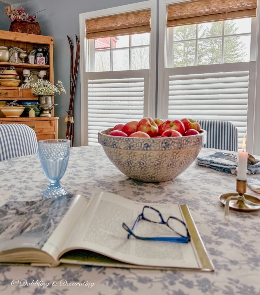 Bowl of apples on blue and white dining room table with three windows with bamboo shades and vintage hutch.