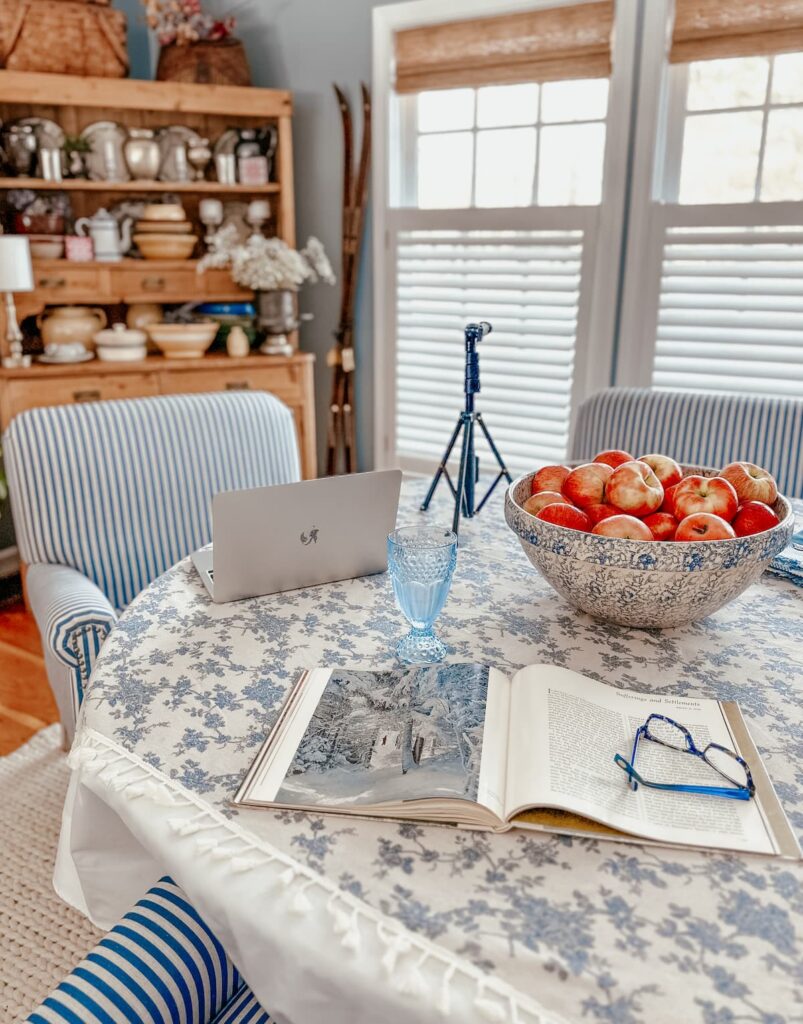 blue and white table with laptop, book, glasses, bowl of apples and tripod in dining room with three large windows working on collaboration.