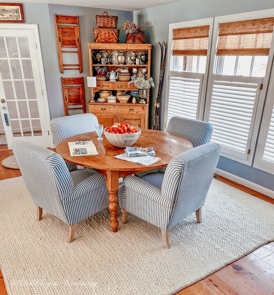 Dining room in an open floor plan with white rug, round wooden table with blue and white striped chairs and three windows with bamboo shades and a vintage hutch.