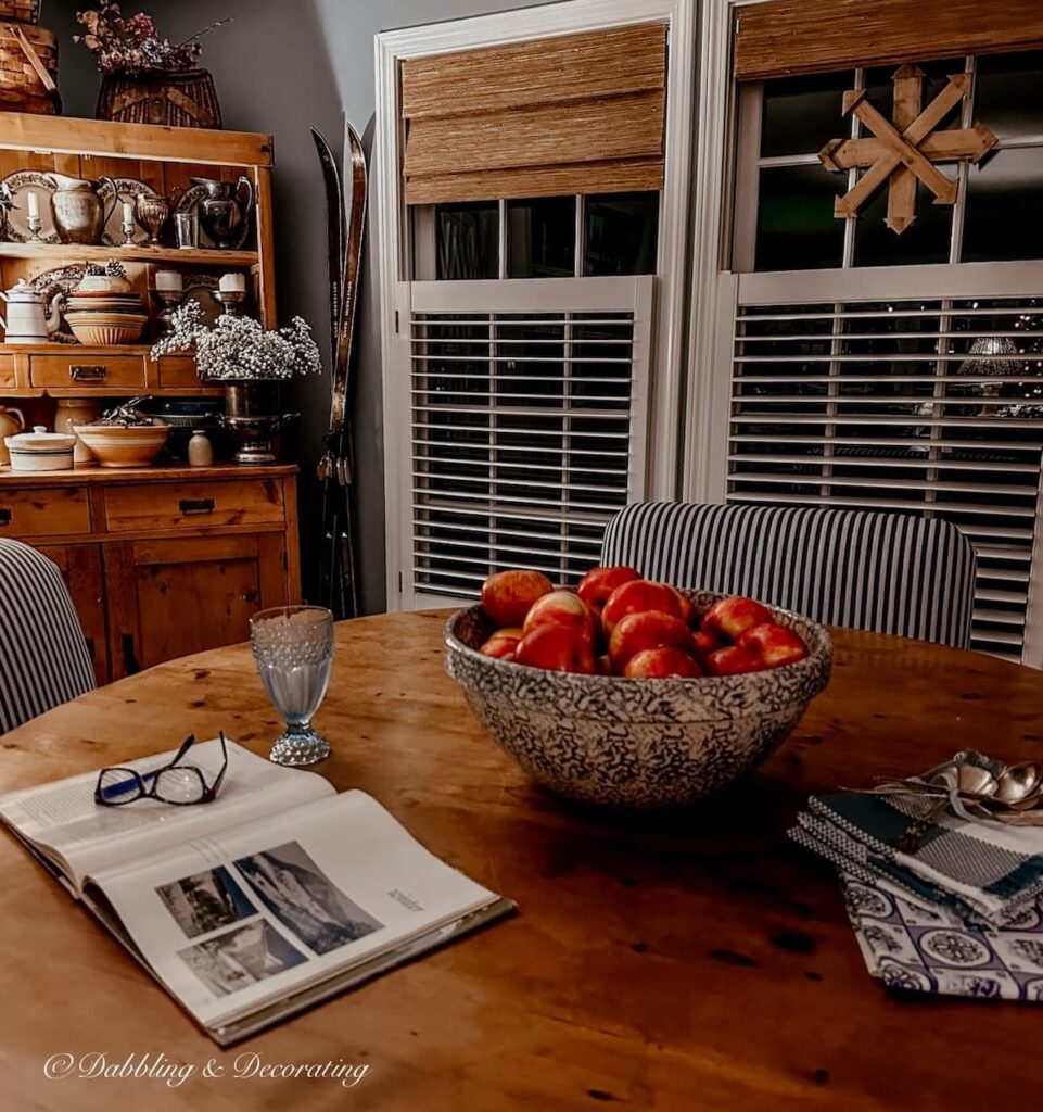 Dining room at night with bowl of apples on the table, open book and light on vintage hutch lit next to windows with TWOPAGE Bamboo Shades.