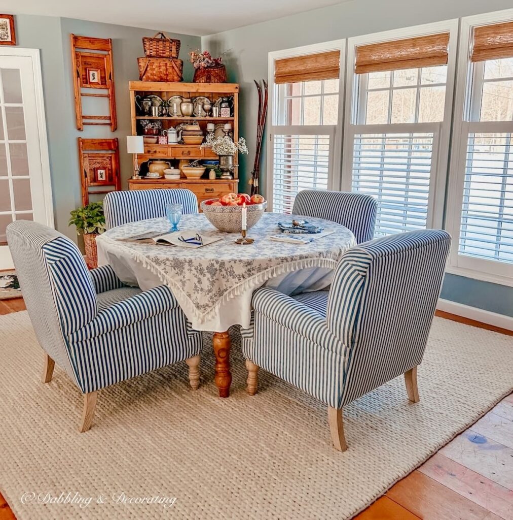 Vintage style dining room with blue and white striped chairs around round table on white rub with vintage hutch and vintage wall decor.