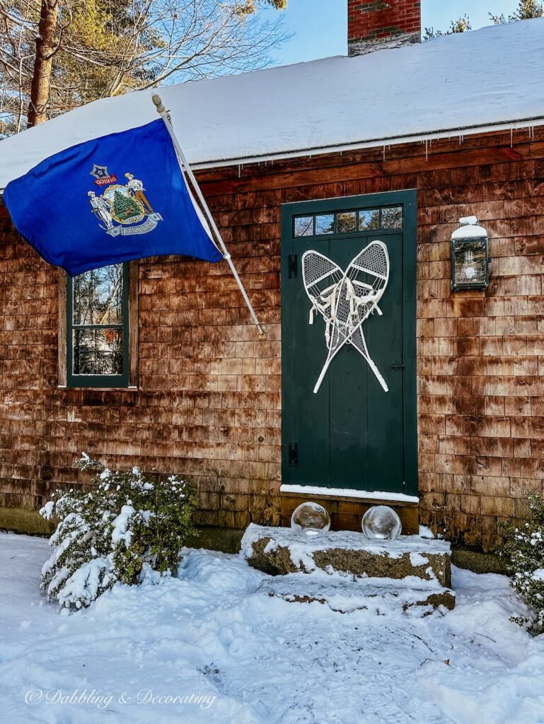 Cedar Shakes house with Maine flag at front door with snowshoes and ice lanterns on snowy steps.