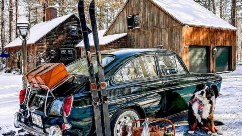 1968 VW Fastback in green with vintage HEAD skis, picnic basket and luggage on trunk with Bernese Mountain Dog in front of cedar shakes home on a snow day.