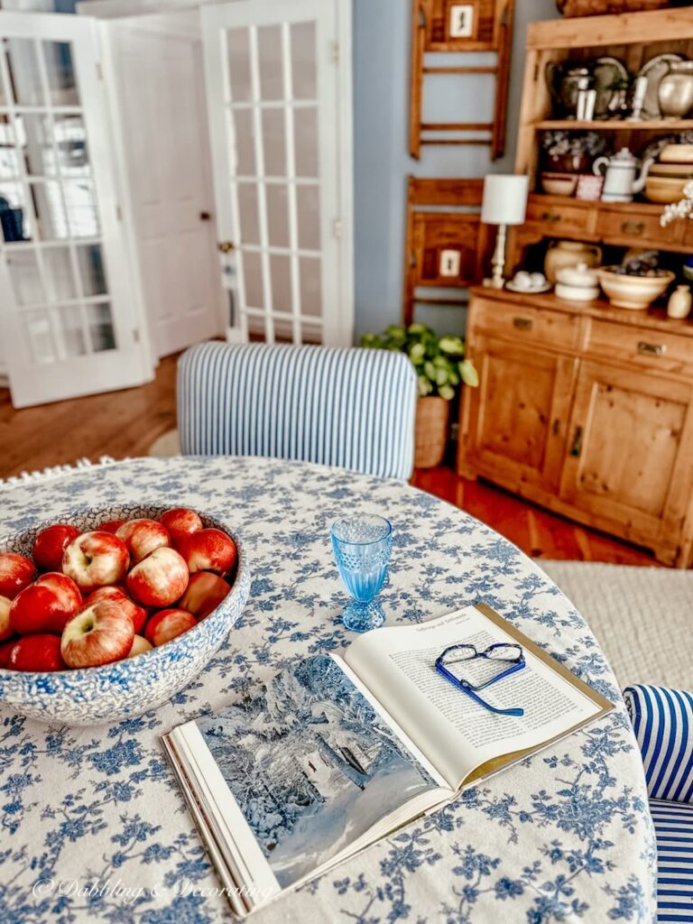 Dining room table with blue table cloth bowl of apples, vintage hutch and vintage wall decor with chairs.