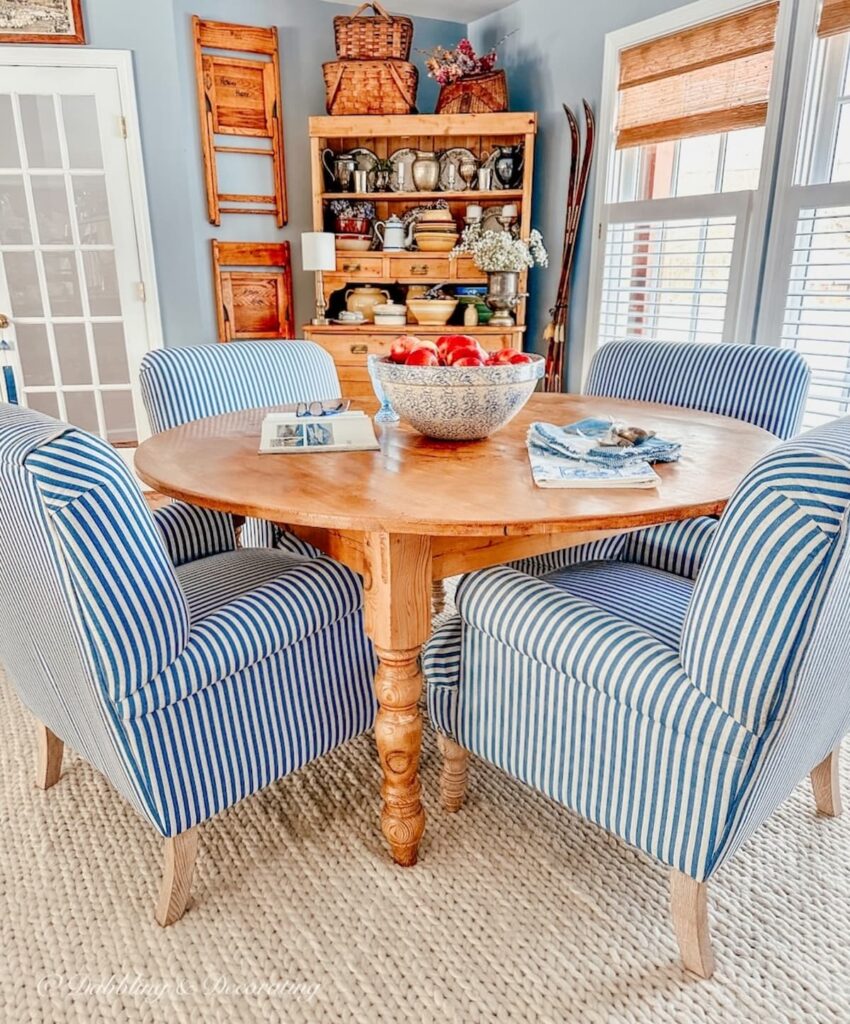 Dining room with round pine table and blue and white striped chairs with vintage hutch and old chairs on wall.