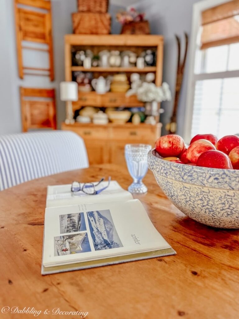 Dining room table with book open and a bowl of apples with vintage hutch and hanging wall chairs in the background.