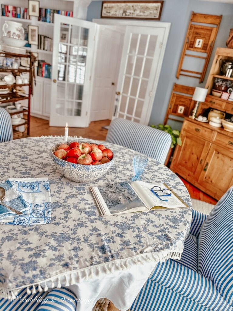 Dining room table with blue table cloth bowl of apples, vintage hutch and vintage wall decor with chairs in Eclectic Interiors.