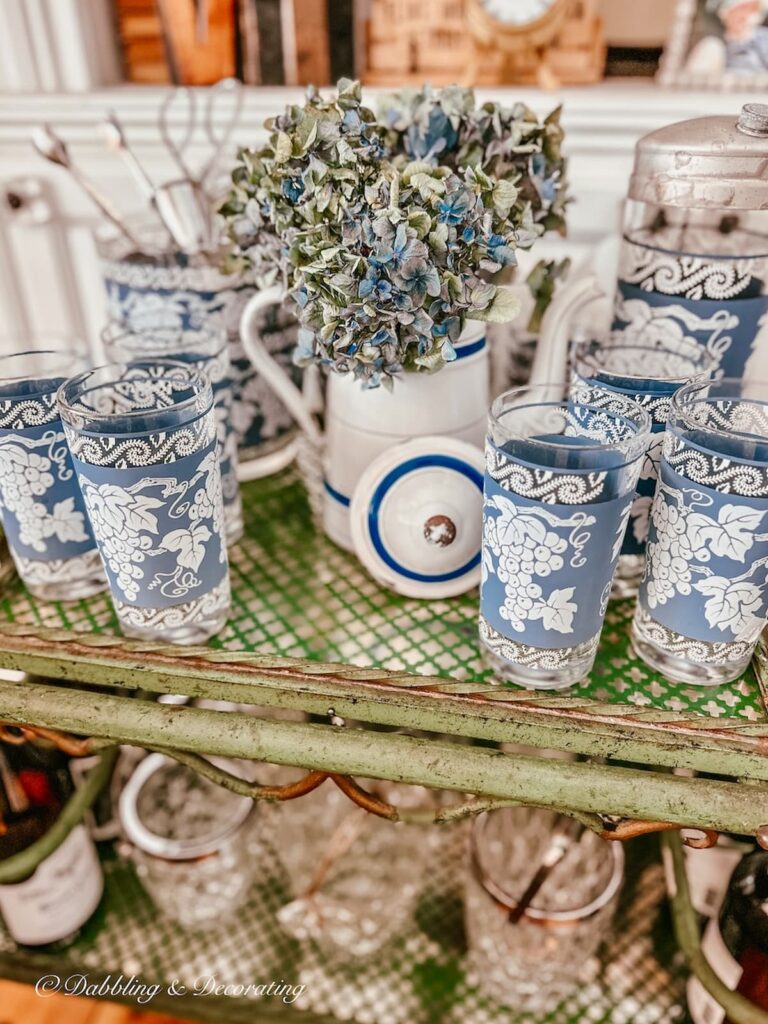 Blue and white Parisian teapot with vintage Jeanette barware on antique bar cart with blue hydrangeas as side table in living room close up.