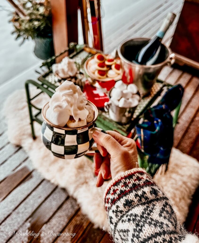 Cup of hot chocolate in hand over antique bar coat decorated for apres' ski on snowy porch.