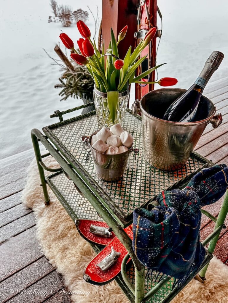 Antique bar cart with marshmallows, ice buckets, and red tulips on snowy winter porch.