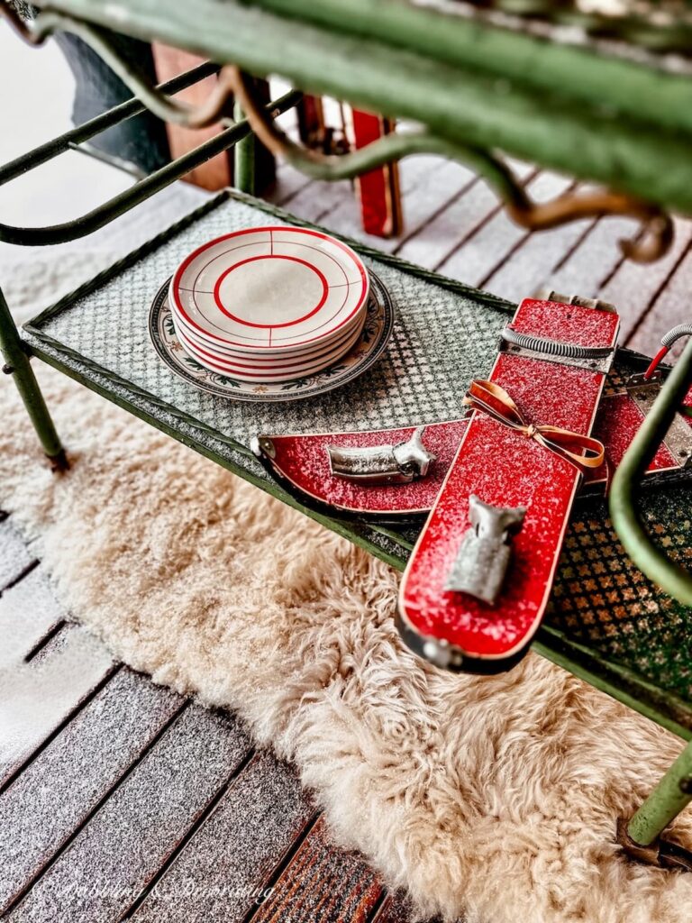 Antique bar cart in green on sheepskin rug on outdoor wintery porch, with red patterned dishes and small skis from Bavaria, Germany.