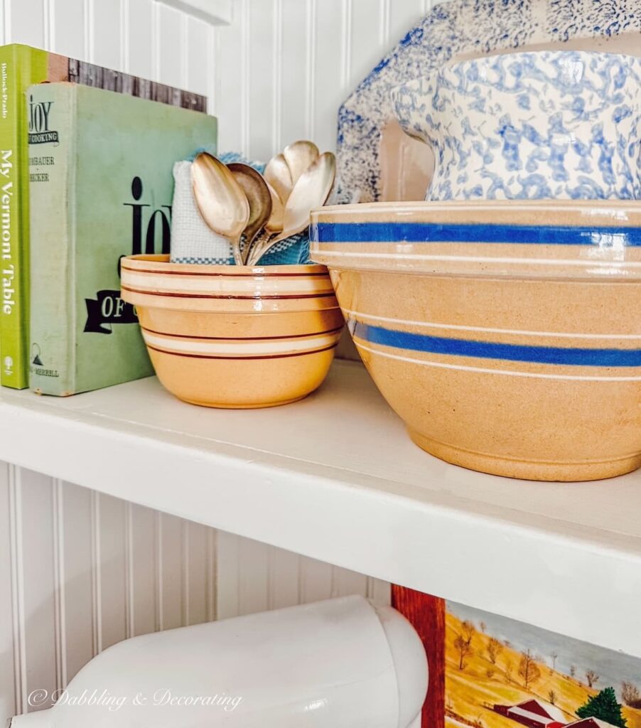 Yellow ware bowls displayed on white kitchen open shelving with two green cookbooks, silver spoons, and blue spongeware pitcher.