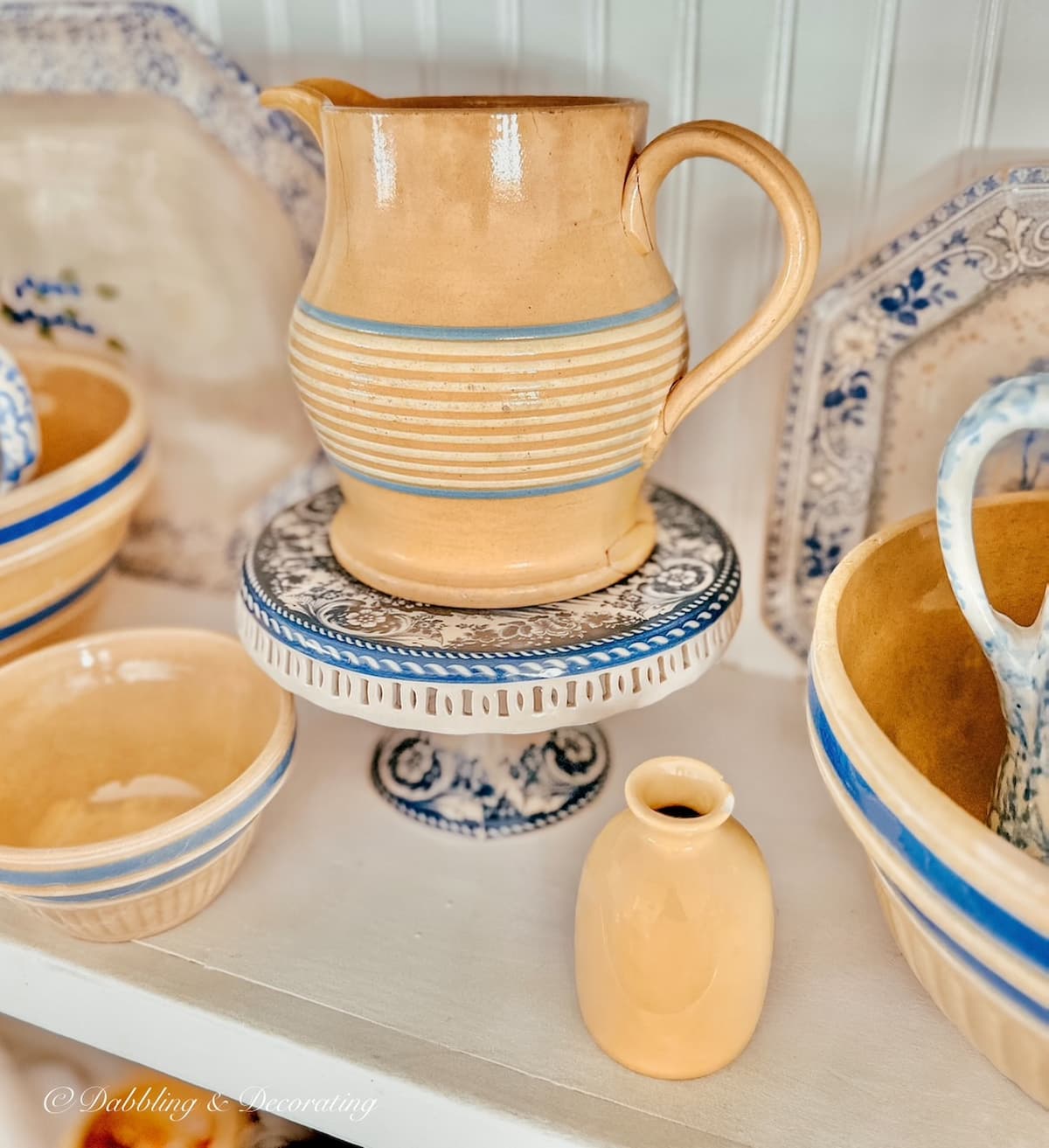 Antique yellow ware pitcher mixed with blue and white cakestand and yellow ware bowl and small jug on white kitchen shelves.