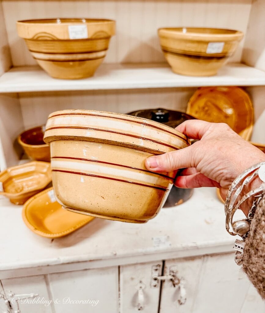 Antique yellow ware with brown and white stripes in hand at antique store in front of hutch filled with vintage yelloware.