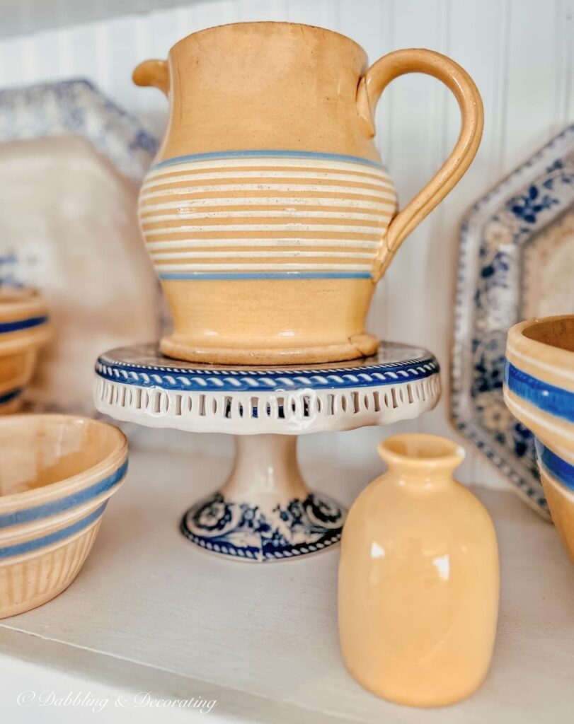 Antique yellow ware pitcher on blue cake stand with vintage yellow ware items on kitchen shelving close up.