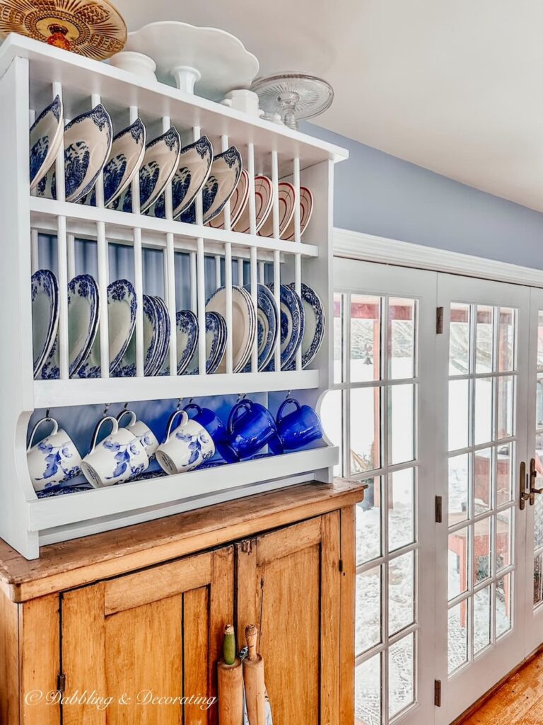 White dish rack filled with blue and white vintage plates and mugs on top of pine wood hutch in kitchen.