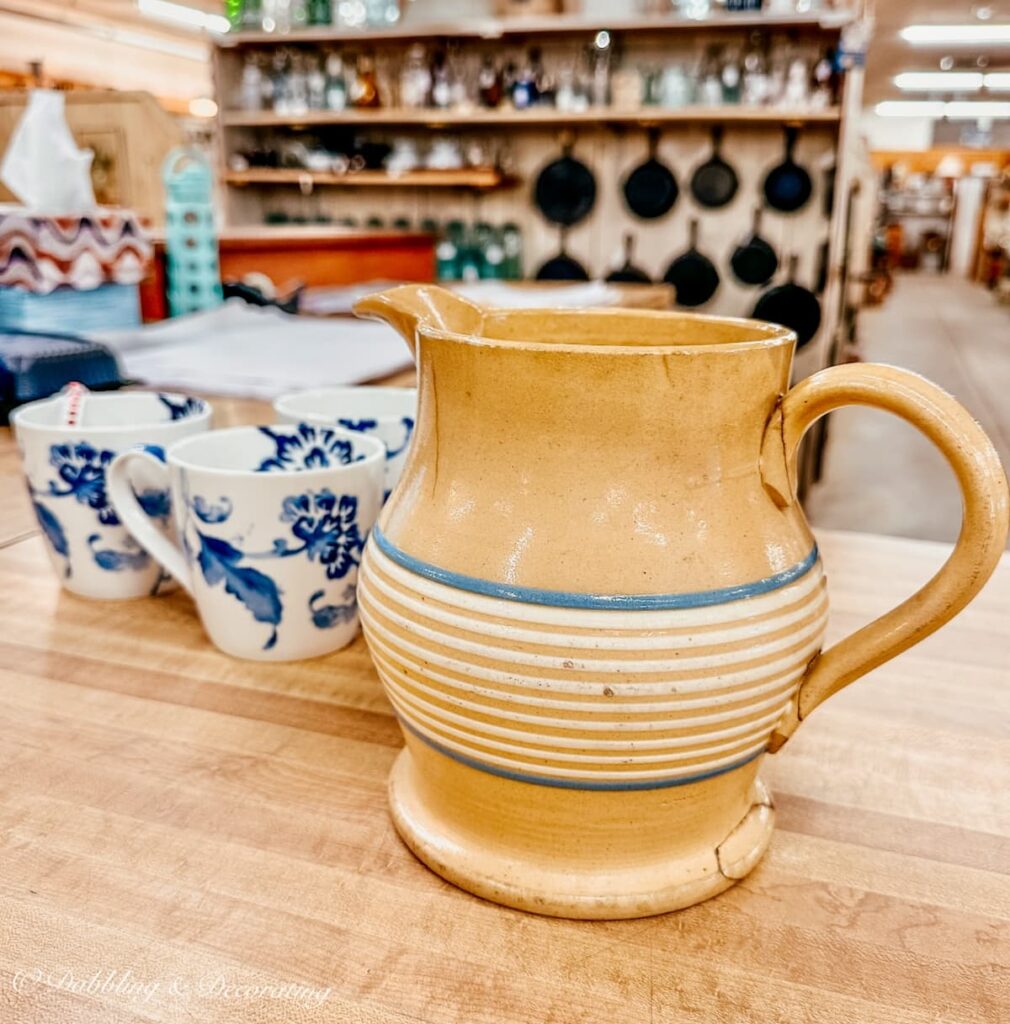 antique yellowware pitcher with blue and white stripes with blue and white mugs on antique store counter for eclectic interiors.